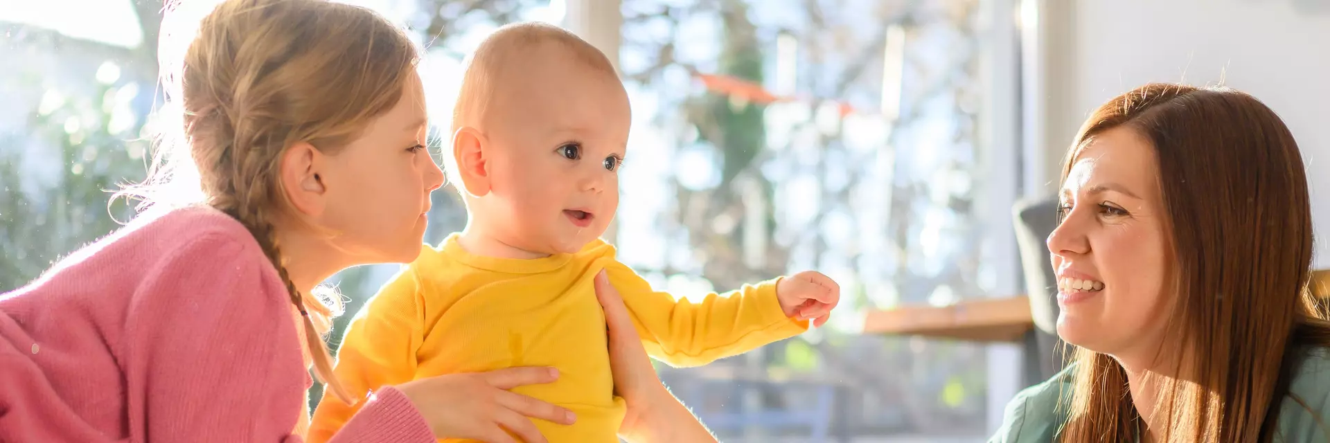 Fröhliches Baby sitzt auf Bett zwischen Schwester und Mama