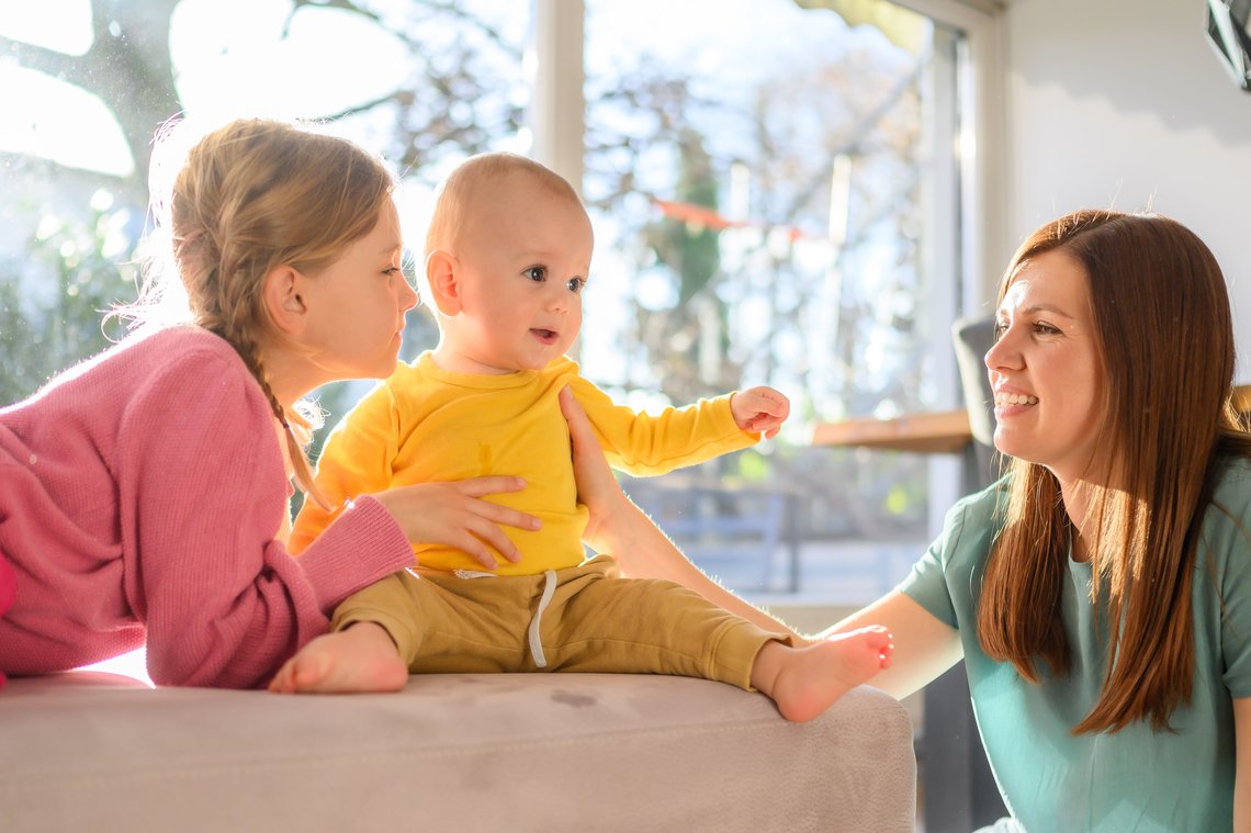 Fröhliches Baby sitzt auf Bett zwischen Schwester und Mama